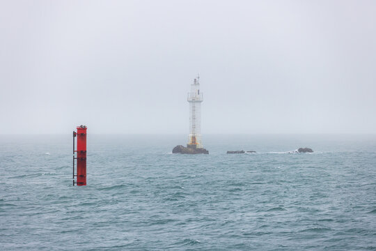 View of a white lighthouse standing on a rocky outcrop amidst choppy sea waves and thick fog, Ushant, Brittany, France.