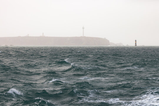 View of rough sea waves with the misty coastline and lighthouses of Ushant island in the distance, Ushant, Brittany, France.