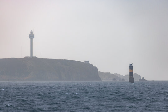 View of a lighthouse on a rocky cliff and a beacon in the choppy sea under a misty sky in Ushant, Brittany, France.