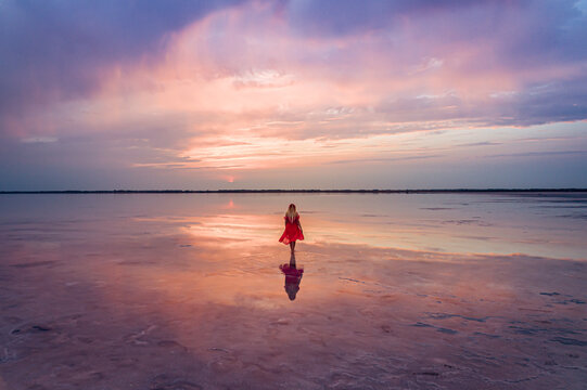 Aerial of a young woman in red dress walking in the water of a unique pink salt lake. Sunset at lake Bursol with beautiful reflections on calm water surface. Stunning scenery