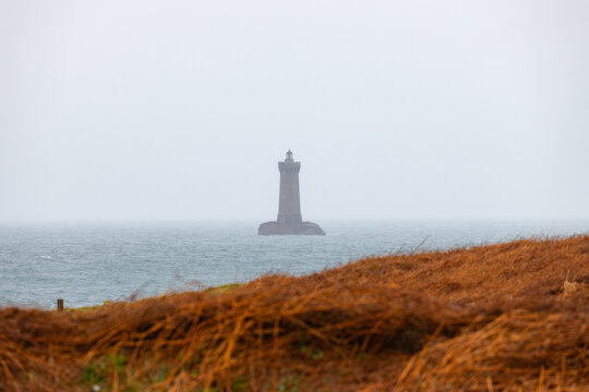 View of Phare du Four lighthouse standing on a rock in the Atlantic Ocean with a foreground of dry orange grass under a misty sky in Porspoder, Brittany, France.