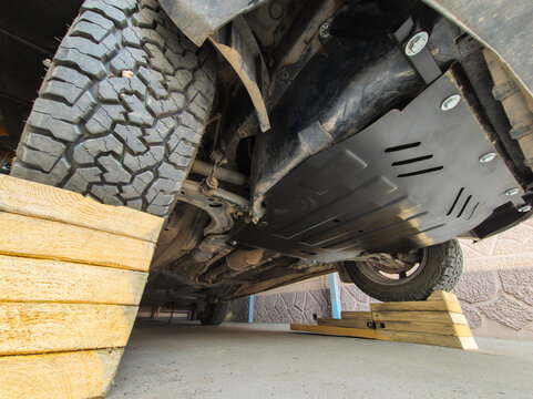 Vehicle underside showcases off-road tires and undercarriage protection plate. Scene set outdoors on wooden ramps during daytime, emphasizing vehicle maintenance.