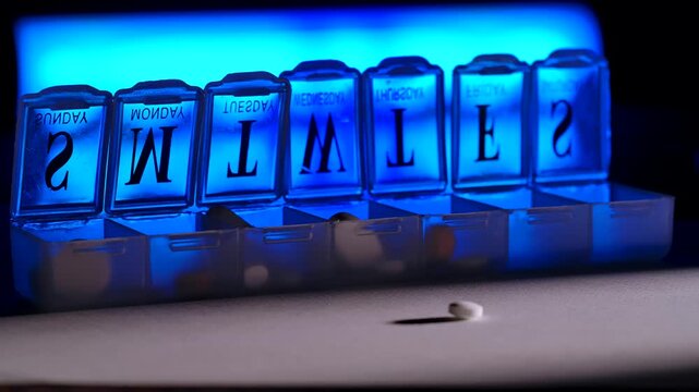 Macro shot of pills falling into a daily pill organizer with labeled compartments in blue light. Concept of healthcare, medication routine, treatment planning, pharmacy and medical advertising.