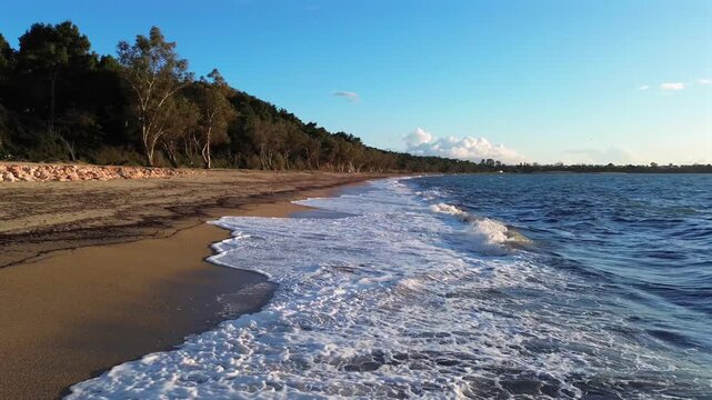 Cinematic drone shot of Monolithi beach in Preveza. High-angle view of sea waves washing onto the long sandy shore, bordered by a lush green forest. Stunning Ionian sea landscape. No audio.