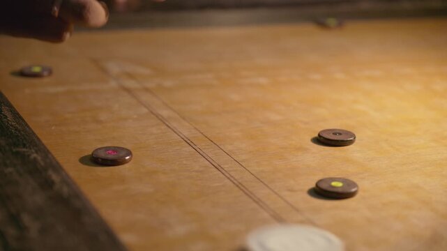 Closeup of player hand striking striker on carrom board, coins moving across wooden surface, dynamic indoor night gameplay showing speed and precision
