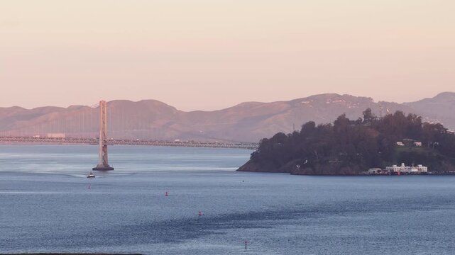 Telephoto shot of a tugboat and barge navigating the calm waters of San Francisco Bay at sunrise, with the Bay Bridge and Yerba Buena Island visible through the morning haze