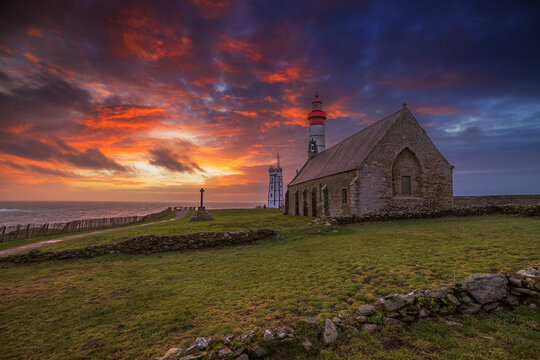 View of the historic Pointe Saint-Mathieu chapel and lighthouse under a dramatic sunset sky in Plougonvelin, Brittany, France.