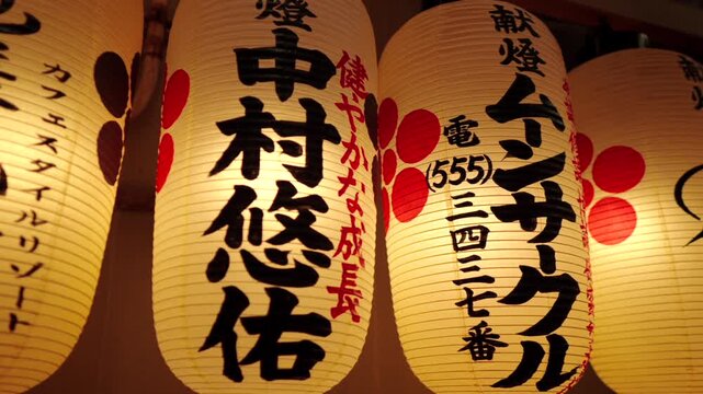 Close-up panning shot of illuminated traditional Japanese lanterns hanging at Nishiki Tenmangu Shrine in Kyoto, Japan, displaying various Kanji characters and designs.
