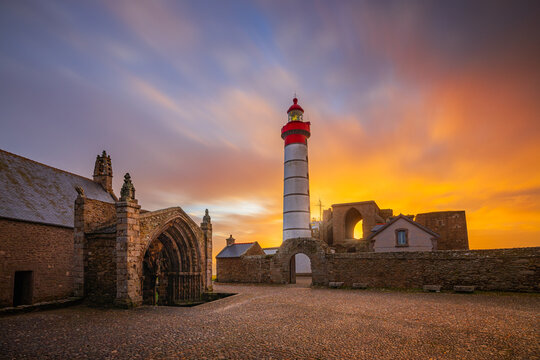 View of Pointe Saint-Mathieu lighthouse and the ruins of the ancient abbey under a vibrant sunset sky in Plougonvelin, Brittany, France.