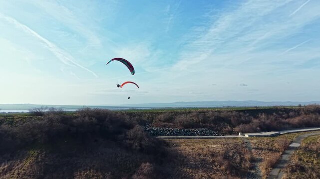 Two people glide through the air in colorful paragliders over a hillside. The scene shows a clear sky and distant mountains on a sunny morning in spring.