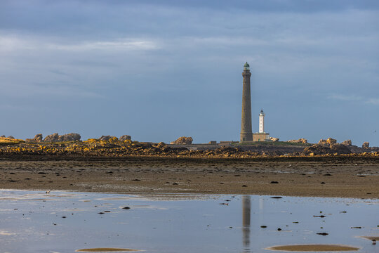 View of Ile Vierge lighthouse standing on a rocky island with its reflection in the wet sand of the beach under a dramatic cloudy sky in Plouguerneau, Bretagne, France.