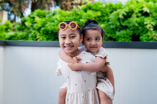 Young girl with round flower sunglasses over her head carrying toddler sister on piggyback