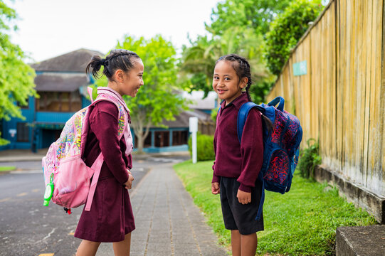 Two young school kids carrying backpacks standing on the sidewalk once facing away