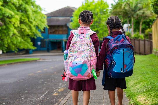 Back profile of two young school kids carrying backpacks while walking on the sidewalk