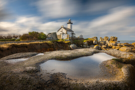 View of Phare de Pontusval lighthouse and its white house standing on a rocky coastline with large boulders and a puddle reflection under a streaky sky in Plouneour-Brignogan-plages, Bretagne, France.