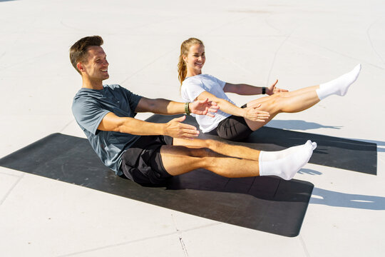 Smiling man and woman doing boat pose core exercise on yoga mats outdoors under bright sunlight