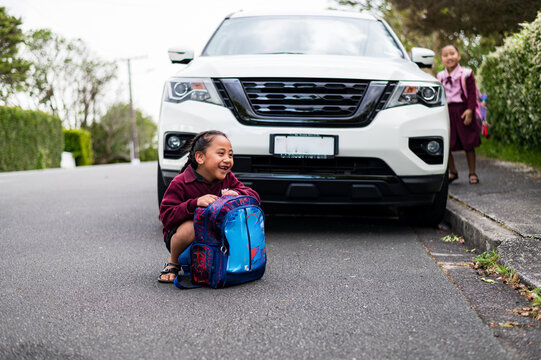 Young boy sits on pavement laughing beside a parked vehicle on the street with sister in background