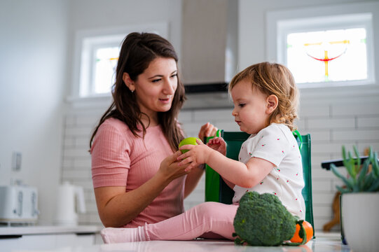 Toddler girl helping mum unpack groceries while seated on kitchen counter