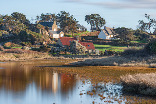 View of traditional stone houses nestled among granite rocks and green trees overlooking a calm coastal inlet in Plougrescant, Brittany, France.