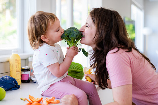 Toddler girl sits on kitchen counter holding a broccoli taking a playful bite with mum