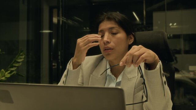 Medium close-up of exhausted Hispanic female corporate executive working to deadline on report computer alone at night in office, grimacing, staring at computer reflecting in glasses