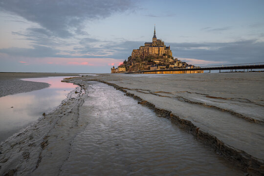 View of Mont Saint-Michel abbey rising above the tidal mudflats with water reflections under a twilight sky in Mont Saint-Michel, Normandy, France.