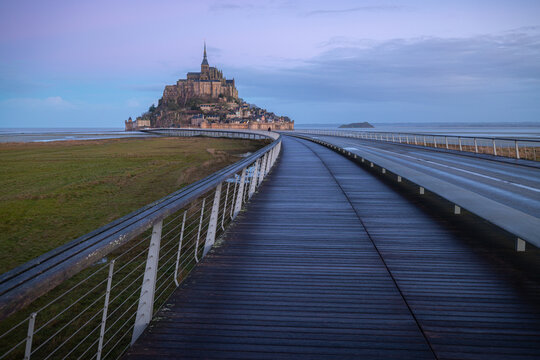 View of Mont Saint-Michel abbey with the modern access bridge, wooden walkway, and surrounding tidal flats under a soft twilight sky Mont Saint-Michel, Normandy, France.