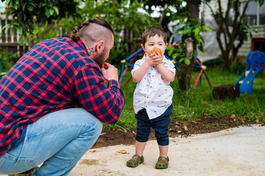 Man in plaid shirt crouching on paved surface in the backyard eating an orange with toddler son