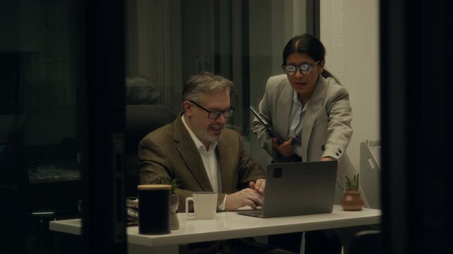 Medium shot of Caucasian male and Hispanic female corporate managers brainstorming together while discussing project, proposing solutions to problems, while working overtime in dark office at midnight