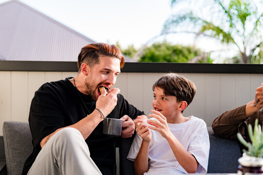 Father taking a big bite while looking at tween son seated on the couch in the balcony