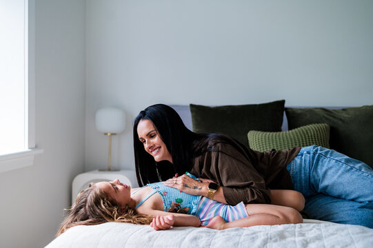 A middle-aged mother and her young daughter play on a bed, smiling and enjoying their time together