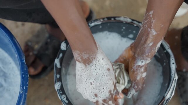 Energetic scrub black hands in bucket, fabric pressed and twisted through thick soap, cleaner parent tackling daily house chores, water splashing over concrete, closeup on palms and foam, sense