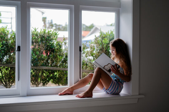 Young girl sitting on a windowsill reading a book inside home