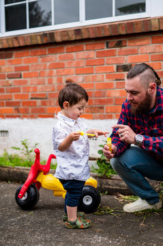 Dad crouching on the paved ground in the backyard playing bubbles with toddler son outdoors