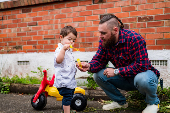 Dad crouching on the paved ground in the backyard playing bubbles with toddler son outdoors