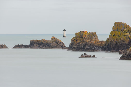 View of the Phare de la Pierre-de-Herpin lighthouse standing in the sea beyond the rocky cliffs of Pointe du Grouin in Cancale, Bretagne, France.