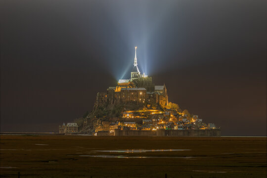 View of the illuminated Mont Saint-Michel abbey at night with a bright spotlight beam from the spire over the tidal flats in Mont Saint-Michel, Normandy, France.