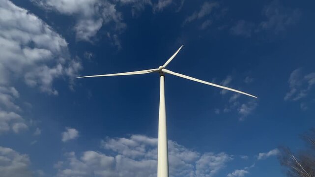 Wind turbine spins against clear blue sky. Clouds drift gently around its white blades. No people appear in this scene. Ideal for renewable energy or environmental visuals