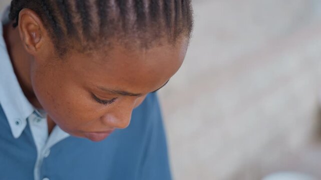 Black woman at house window focused on small repair during chores, leaning over hands threading needle, braided cornrows, blue collared shirt, closeup detail of concentrated expression, quiet evening