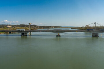 Fototapeta premium Montfaucon, France - 03 19 2026: View of a motorway bridge above Rhône river with trucks driving, an open air career and hills behind in sunny spring blue sky