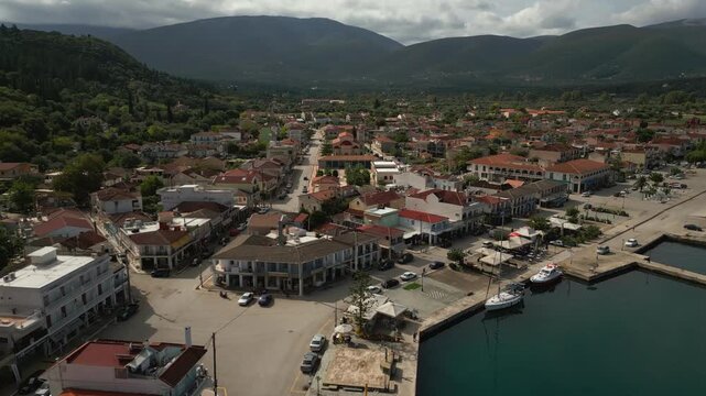 An aerial view of a coastal town with traditional buildings and a harbor, nestled against a lush green mountainside under a cloudy sky.