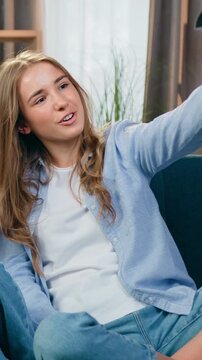 Good-looking smiling satisfied loving blond mother sitting on soft couch in cozy sitting room together with eldest and younger daughters during pleasant video chat on smartphone,slow motion