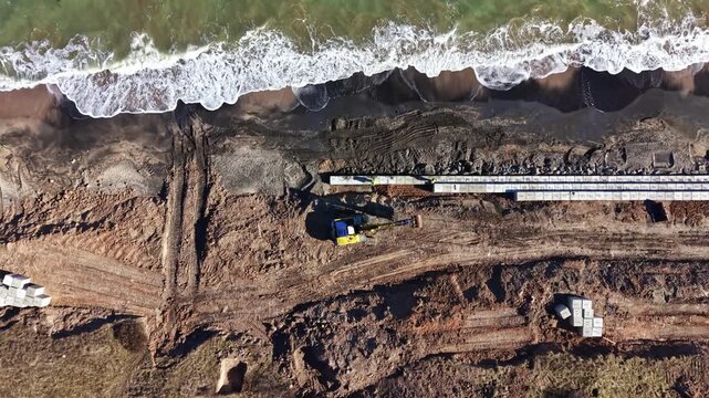 Heavy machinery works near the beach where the ocean meets the land. Rows of materials are placed along the shore in a construction area.