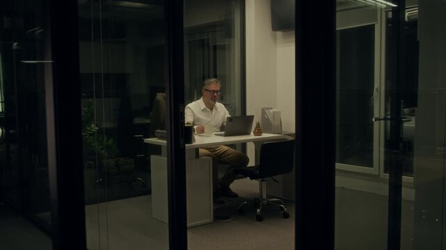 Wide shot of mature Caucasian male manager sitting at desk, typing on laptop, finishing work, picking up jacket and leaving office to go home in middle of night, switching off light