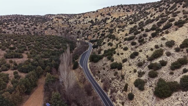 A winding road leads through rocky hills and patches of greenery in a remote area. The sky is overcast, creating a muted atmosphere. The landscape shows a mix of terrain and foliage.