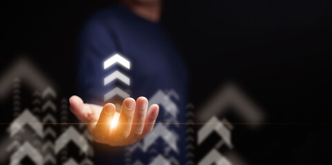 Hand holding glowing upward arrows for business growth, profit, and career success. Abstract digital concept for leadership, progress, and innovation with copy space on dark background for text. © OleCNX