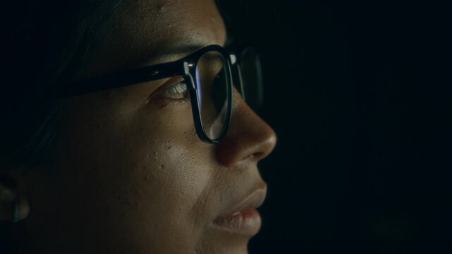 Side extreme close-up of face of Hispanic woman in eyeglasses attentively studying and analyzing business data on computer screen while working at midnight in dark office to deadline