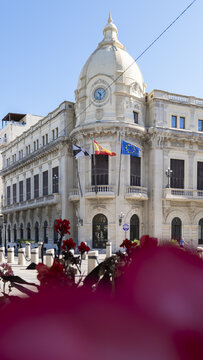 View of the historic Palacio de la Asamblea building with its white facade, dome, and clock tower behind red flowers Ceuta, Ceuta, Spain.