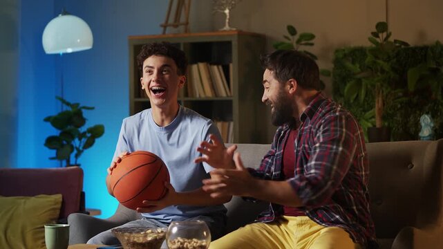 Teen boy and his father watching basketball game on TV, happy with team win