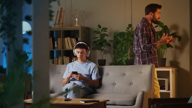 Displeased father scolding rebellious teen son in headset playing on phone